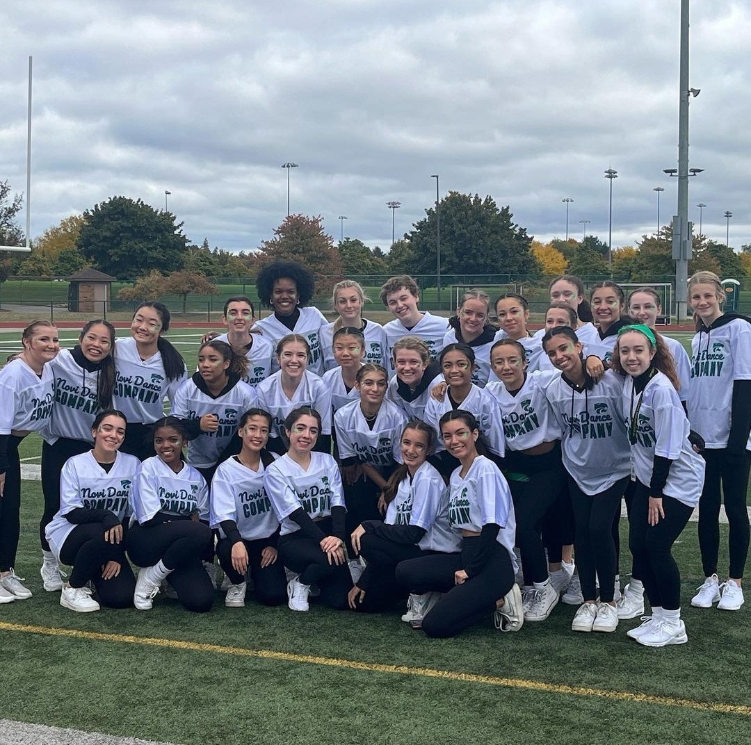 A group of high school dance students in white football jerseys smiling in a group on a football field