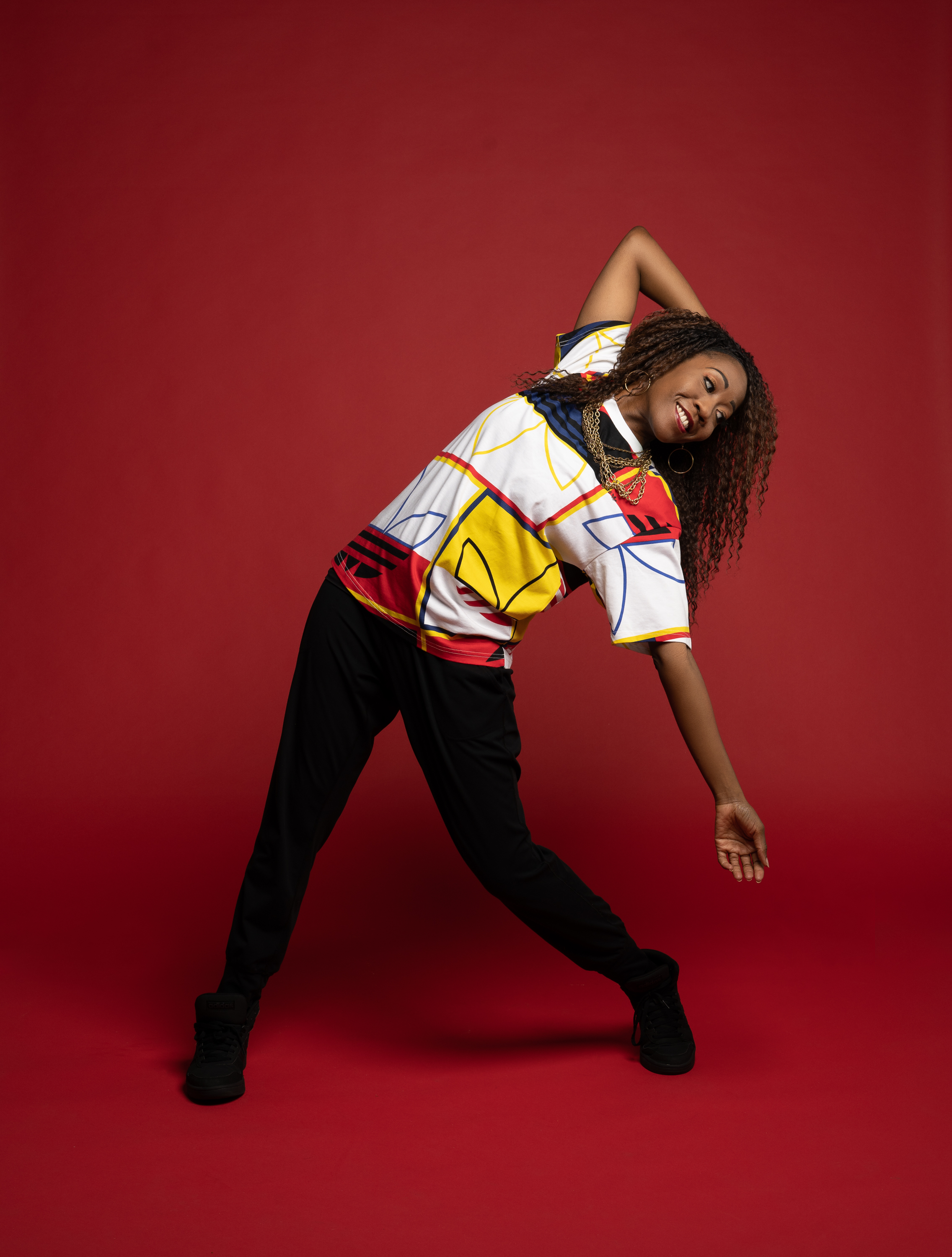 A black woman posing with her hand on her head and leaning to the right, wearing an absract colorful top and black pants against a red back round.